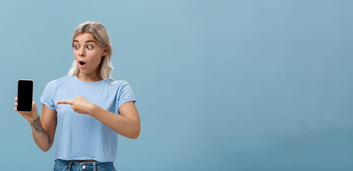 Girl being amazed with cool new smartphone received on birthday. Surprised speechless happy woman with fair hair and tattoo dropping jaw holding smartphone pointing and staring at gadget screen.