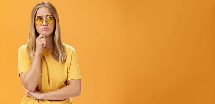 Troubled and concerned young stylish woman facing troublesome problem thinking holding hand on chin pursing lips looking at upper right corner thoughtfully standing against orange background. Lifestyle.