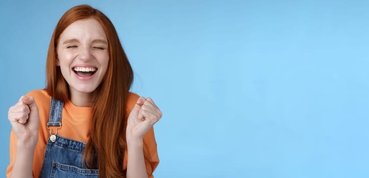 Lifestyle. Sincere happy rejocing ginger girl close eyes smiling broadly say yes waving clenched fists joyfully celebrate enterting university dream come true winning prize triumphing cheerfully blue background.