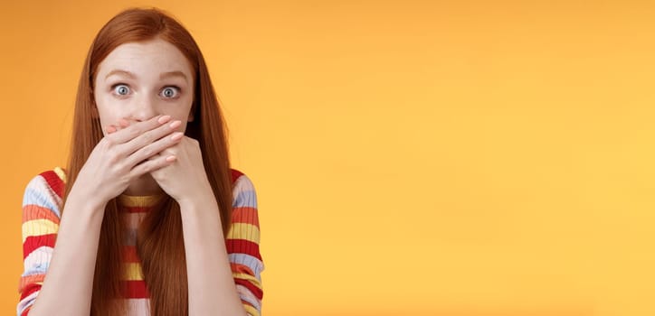 Oops sorry. Girl feeling awkward saying inappropriate word standing unconfident cover mouth palms wide eyes staring camera shocked express surprise nervously standing orange background.