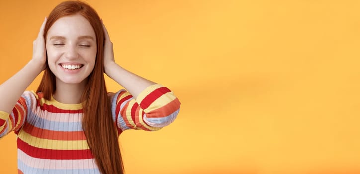 Dreamy charismatic relaxed tender redhead caucasian girl 20s close eyes cover ears imaging far away, smiling broadly enjoy silence peace comfort without sounds noise-free, orange background.