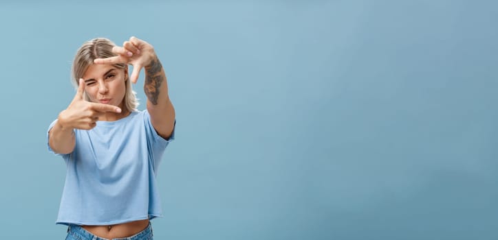 Artistic and creative female designer in stylish t-shirt closing one eye folding lip and making frame gesture while looking through it as if taking measurement or picturing something over blue wall. Copy space