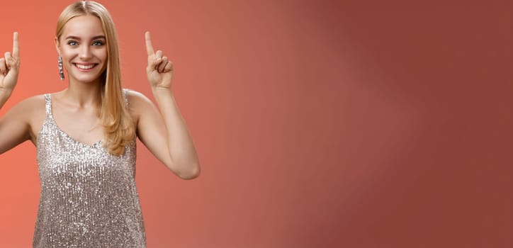 Excited gorgeous blond european woman in silver shiny elegant dress raise hands pointing up showing impressive incredible advertisement smiling happily thrilled wanna take closer look, red background.