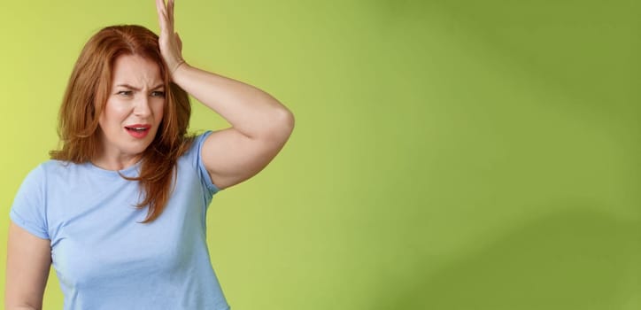 Something important slip my mind. Concerned worried upset redhead mature woman punch forehead turn away frustrated frowning disappointed forget cancel appointment stand green background.