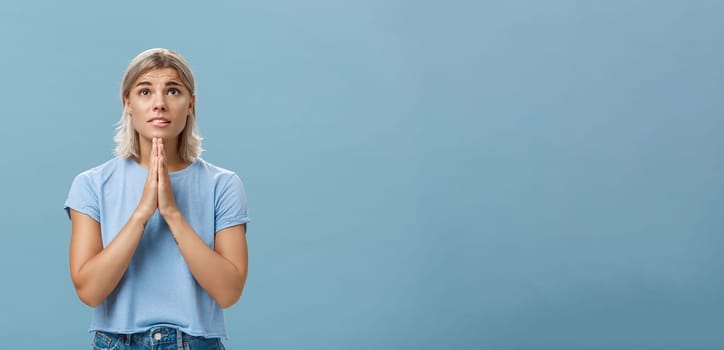 Studio shot of hopeful focused dreamy girl with attractive face and blonde hair holding hands in pray near body looking up hopefully with faith praying or making wish over blue background. Body language concept