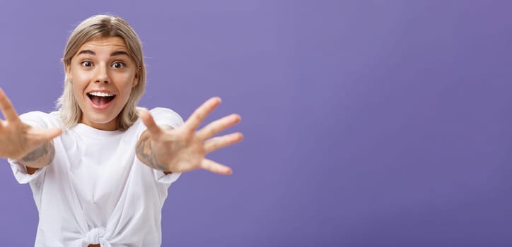 Lifestyle. Waist-up shot of amused and excited attractive stylish young woman in white t-shirt pulling hands at camera with desire smiling thrilled and happy wanting hug or take something over purple background.