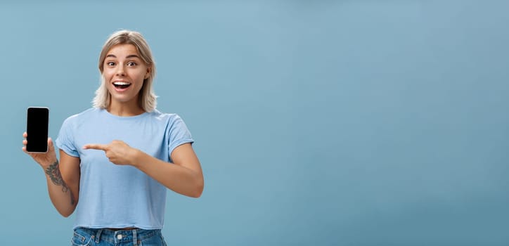 Lifestyle. Waist-up shot of thrilled and impressed good-looking female student in casual t-shirt smiling joyfully pointing at smartphone screen showing awesome place via internet to friend over blue background.