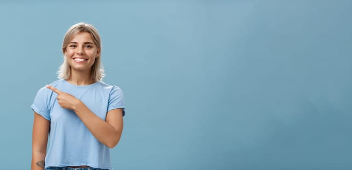 Come with me. Portrait of stylish pleased good-looking female student with blond hair in trendy t-shirt pointing at upper left corner and smiling with delight and happiness posing over blue background.