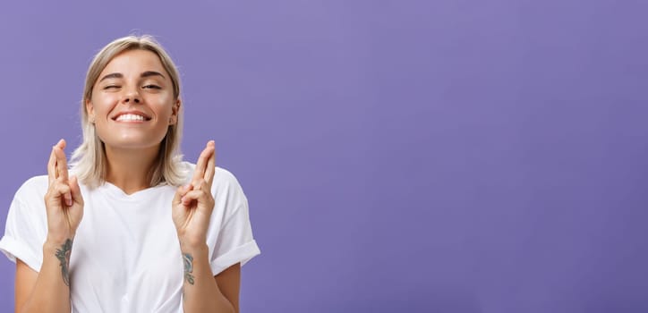 Waist-up shot of hopeful optimistic attractive stylish woman in white t-shirt with tattooed arms winking smiling joyfully while crossing fingers for good luck making wish over purple background. Body language concept