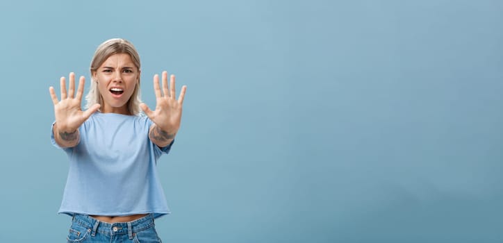 Hold right there. Portrait of intense displeased and irritated attractive young female in blue t-shirt pulling hands towards camera in stop or not gesture frowning and making annoyed expression.