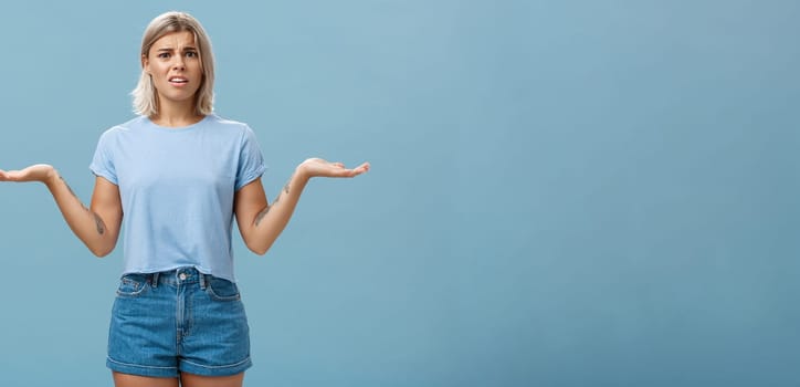 Girl feeling struggles to understand what happening. Confused perplexed intense good-looking female with fair hair shrugging with hands spread aside in displeased clueless pose posing over blue wall.