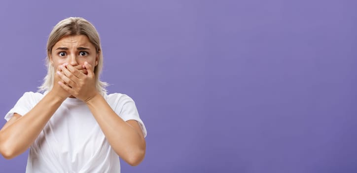 Waist-up shot of shocked nervous and scared young woman witnessing terrible crime covering mouth with both hands not to scream frowning staring frightened at camera over purple background. Emotions concept