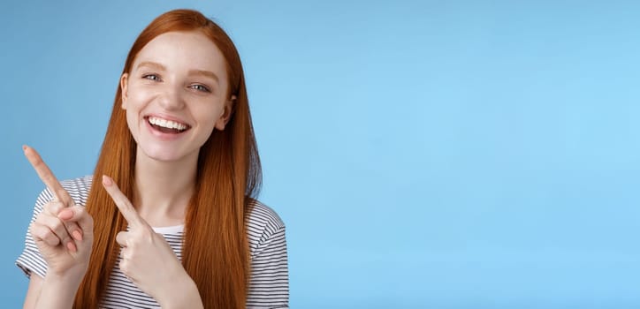 Happy carefree smiling redhead caucasian girl ginger straight hairstyle pointing upper left corner laughing joyfully amused cool promo giving recommendation try new product, standing blue background.