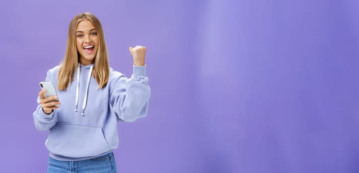 Yes we did it. Pleased excited woman in hoodie raising fist in celebration and success gesture smiling broadly triumphing holding smartphone reacting to positive news in cellphone over purple wall. Technology and emotions concept