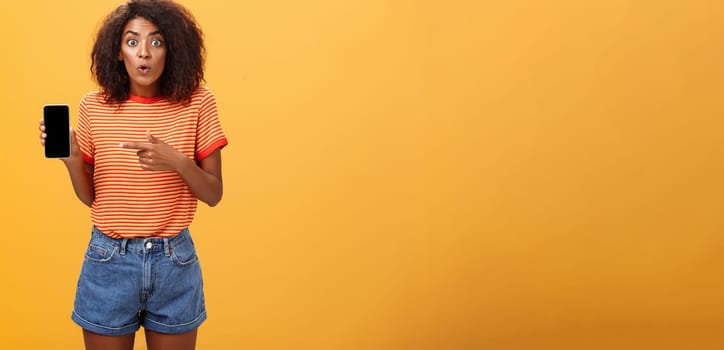 Portrait of impressed and curious female friend showing profile of ex to friend talking about shocking changes of appearance folding lips from interest holding smartphone pointing at cellphone screen. Lifestyle.