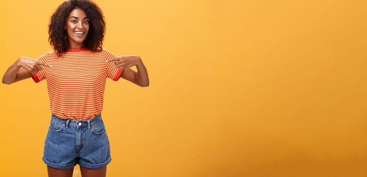 Hey pick me I your girl looking for. Portrait of charming friendly-looking ambitious dark-skinned female with afro hairstyle pointing at chest proudly and joyful posing against orange background.