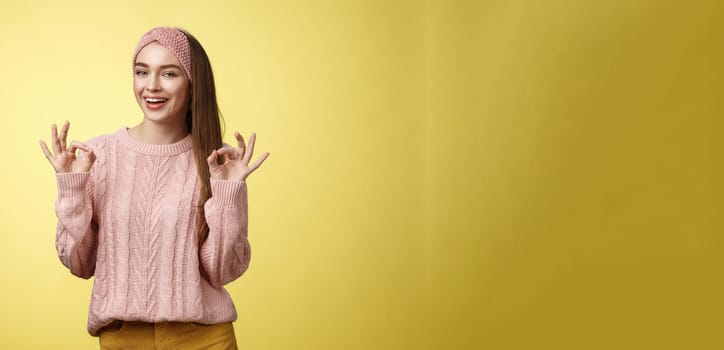 Positive attractive upbeat young cute european woman in 20s wearing casual sweater, headband showing okay, ok gesture smiling assuring everything, good, fine smiling confident over yellow wall.