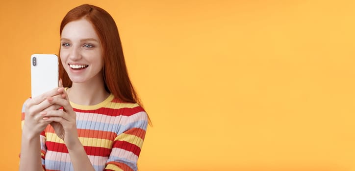 Redhead girl having fun recording hilarious friend actions hold smartphone look display amused shooting funny video telephone standing orange background satisfied smiling delighted. Lifestyle
