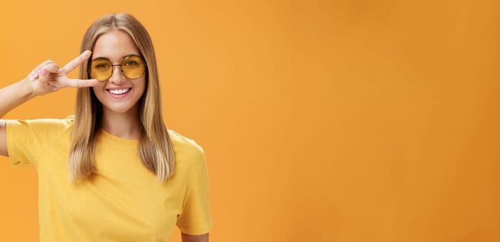 Cute optimistic and friendly young caucasian woman with fair hair in yellow t-shirt and sunglasses showing peace sign near eye and smiling cheerfully at camera taking shot over orange background. Lifestyle.