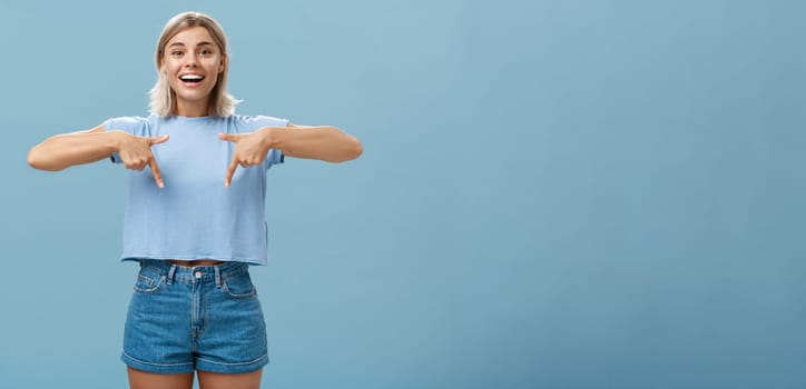 You have to see this. Portrait of joyful amused and happy stylish blonde female in trendy t-shirt pointing down with arms near chest smiling broadly showing amazing copy space over blue background.
