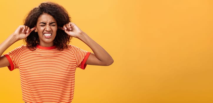 Waist-up shot of intense bothered dark-skinned woman clenchign teeth from discomfort and antipathy closing ears not hear botherind loud noise standing displeased and annoyed over orange wall. Copy space