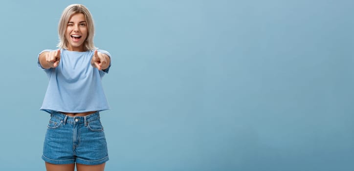 Happy enthusiastic stylish blonde female with tattoos pointing at camera as if picking or making hint smiling joyfully and winking from amazement and happiness posing over blue background. Copy space