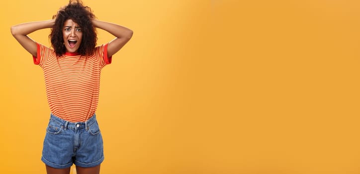 Girl panicking not being ready meet boyfriend parents. Portrait of shocked troubled and concerned gloomy girlfriend with afro hairstyle holding hands on hair yelling from shock over orange background. Copy space