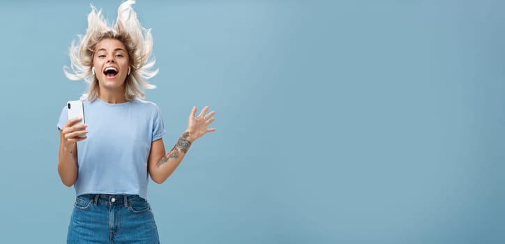 Expressing happiness with help of great tunes. Joyful amused and happy good-looking young female student jumping having fun listenign music in wireless earbuds, holding smartphone over blue wall.