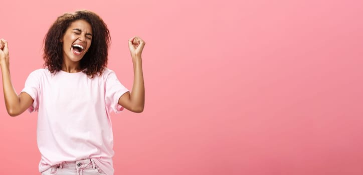 Amazed and happy triumphing african american sportswoman celebrating victory yelling from awesome exciting feelings closing eyes raising fists hight in win gesture standing over pink background. Lifestyle.