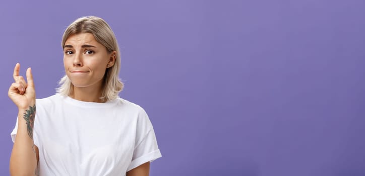 Girl having tiny problem. Concerned attractive blonde girl with tattoo on arm pursing lips in troubled look shaping small or little object, dissatisfied with regret in eyes over purple wall. Copy space