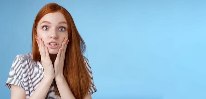 Shit bummer. Shocked worried young intense serious-looking confused young redhead girl look stressed nervously touching face wide eyes staring camera anxiously, standing blue background.