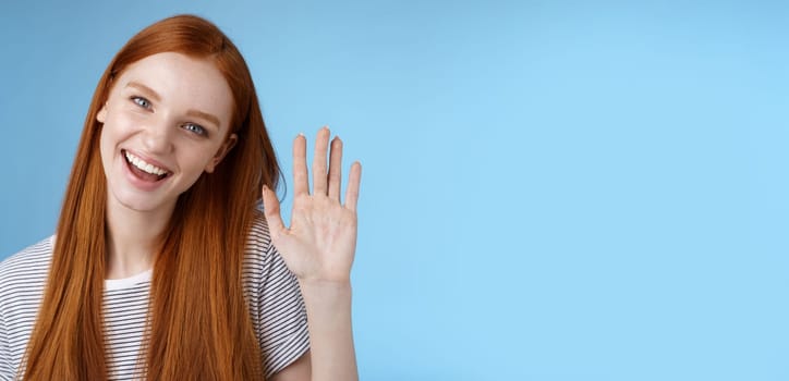 Attractive confident redhead sassy girl pure clean skin blue eyes tilting head cheerfully waving hand hello hi gesture greeting you look camera friendly welcoming friend, standing studio background.