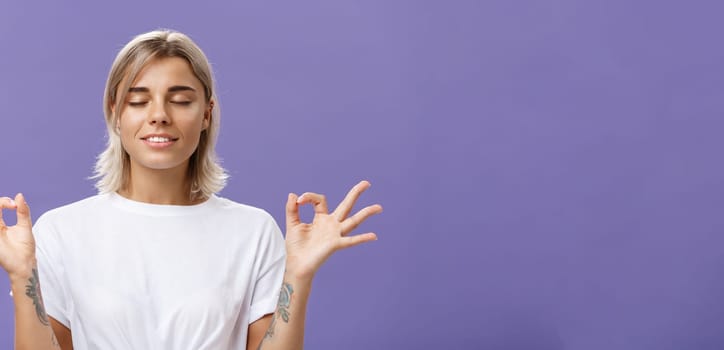 Peaceful calm good-looking female blonde in white t-shirt closing eyes smiling relieved and happy feeling satisfied with life standing in lotus pose with zen gesture over purple background. Copy space
