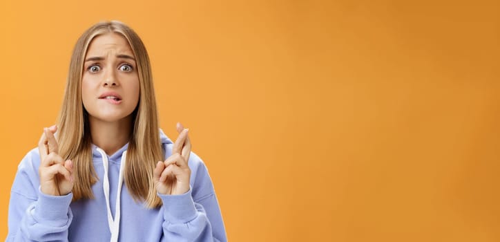 Worried anxious hopeful young woman with fair hair in stylish over-sized hoodie biting lower lip frowning concerned crossing fingers for good luck praying for dream come true against orange wall. Lifestyle.