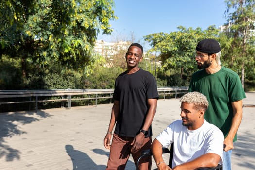 Young disabled Black man in a wheelchair and his male friends walking together in the city. Copy space. Friendship concept.
