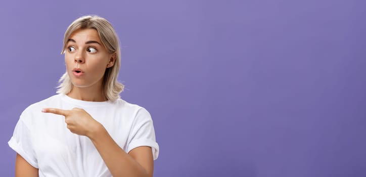 Close-up shot of intrigued interested attractive adult blonde female in white t-shirt folding lips staring and pointing left with curious and thrilled expression over purple background. Advertisement concept