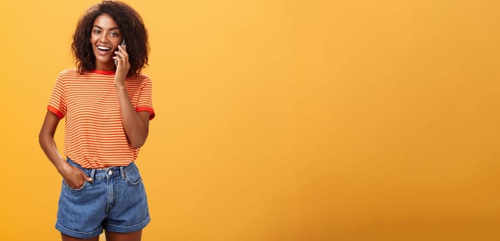 Indoor shot of stylish confident female coworker in trendy t-shirt and denim shorts holding smartphone near ear calling friend talking via cellphone casually about girls stuff over orange background. Lifestyle.