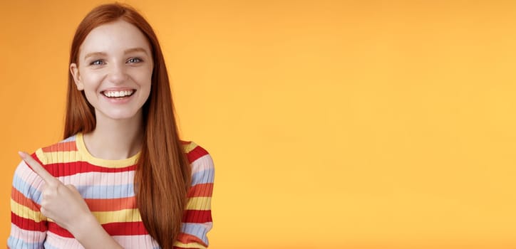Happy charismatic smiling young ginger girl blue eyes sincere tender grin laughing broadly having fun discuss hilarious concert pointing upper left corner showing awesome product, orange background.