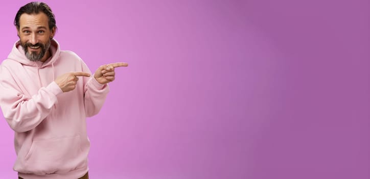 Sassy confident good-looking macho man 50s in pink hoodie smiling broadly inviting join pointing right showing interesting amusing place hang out welcoming take look, standing purple background.