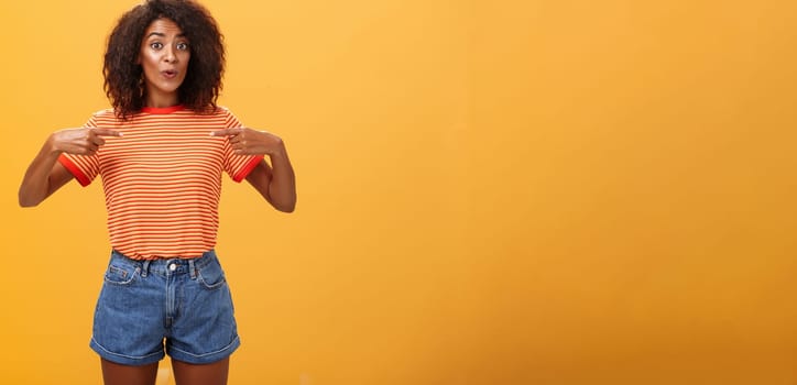 Woman being delighted telling friend she got new job pointing at herself with pleased satisfied look bragging standing happy over orange background in stylish denim shorts and striped t-shirt. Lifestyle.