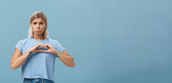 Heart being broken. Sad and gloomy heartbroken girl with blond hair tattoos on arms and tanned skin pursing lips whining and complaining making love sign over breast standing unhappy near blue wall.