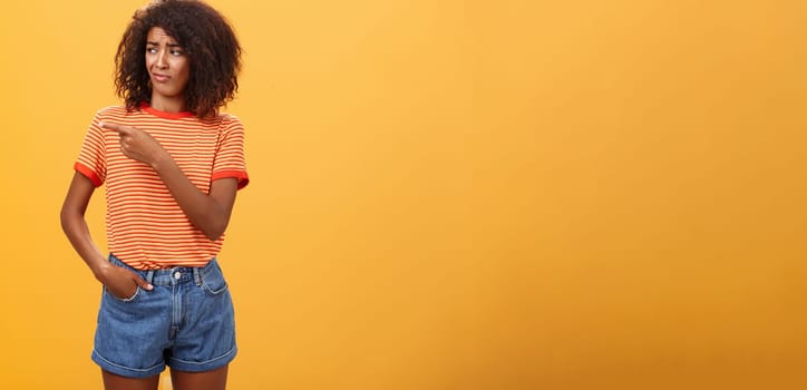 Girl looking at doubtful unimpressive perfomance of model. Portrait of displeased confused good-looking dark-skinned female with afro hairstyle looking and pointing left with scorn and indifference.