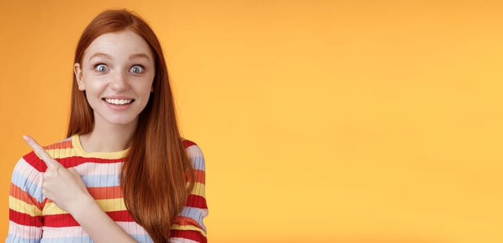 Excited thrilled good-looking young silly redhead girl surprised pop eyes camera gasping impressed pointing upper left corner see super star grinning excitement delight, orange background. Copy space