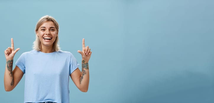 Waist-up shot of upbeat creative stylish and gorgeous blonde female student have tattoos on arms pointing up with raised forefingers smiling with delight and joy posing over blue background. Advertisement concept