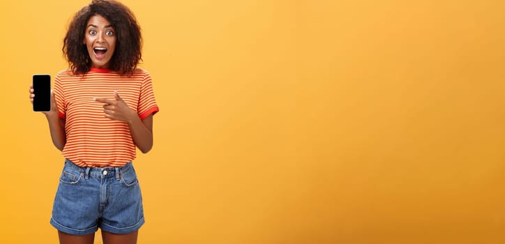 Impressed fascinated stylish slim african american curly-haired woman in striped t-shirt dropping jaw from amazement holding awesome smartphone pointing at device screen over orange wall. Lifestyle.