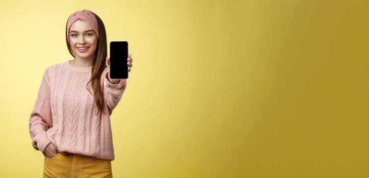 Best smartphone here you go. Charming outgoing young pretty woman in knitted sweater, headband extending hand with phone showing gadget screen smiling recommending cellphone over yellow wall.