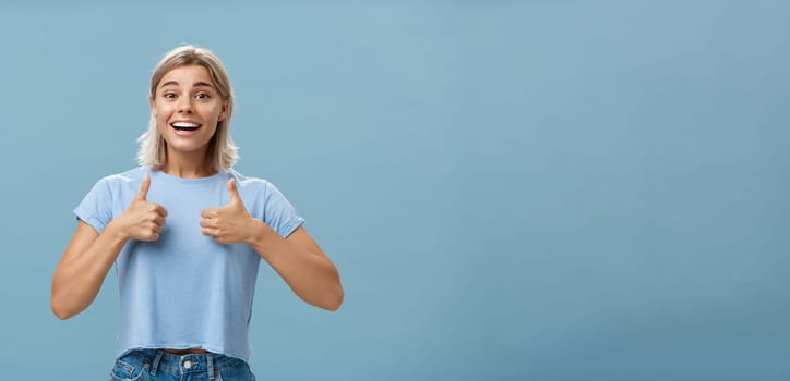 You did great proud of you. Portrait of satisfied and fascinated attractive happy girlfriend in casual t-shirt showing thumbs up and smiling broadly being supportive and cheerful over blue wall.