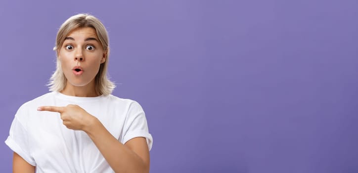 Waist-up shot of overwhelmed excited and surprised attractive urban female in white t-shirt folding lips gasping from amazement being questioned asking question and pointing left over purple wall. Copy space
