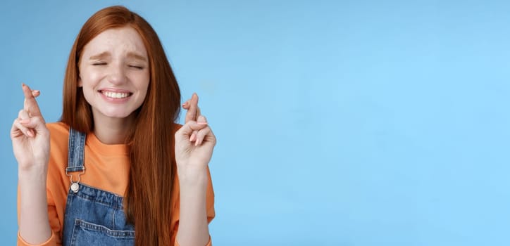 Sincere hopeful cute redhead girl believe miracle close eyes faithfully praying cross fingers good luck hope dream come true make wish eager hear positive results, anticipating blue background.