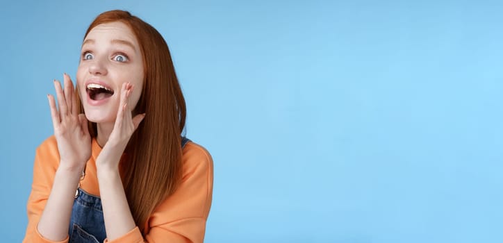 Attractive silly european redhead young girl 20s calling friend searching someone crowd look relaxed joyfully yelling hold hands opened mouth shouting name louder look left, blue background.